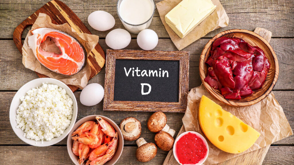 Assorted foods rich in vitamin D—salmon, eggs, cheese, shrimp, liver, mushrooms, milk, butter, cottage cheese—surround a chalkboard labeled “Vitamin D.”