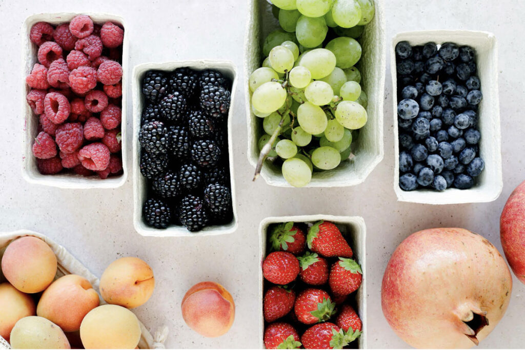 Assorted fresh fruits in containers including berries, grapes, peaches, and pomegranate.