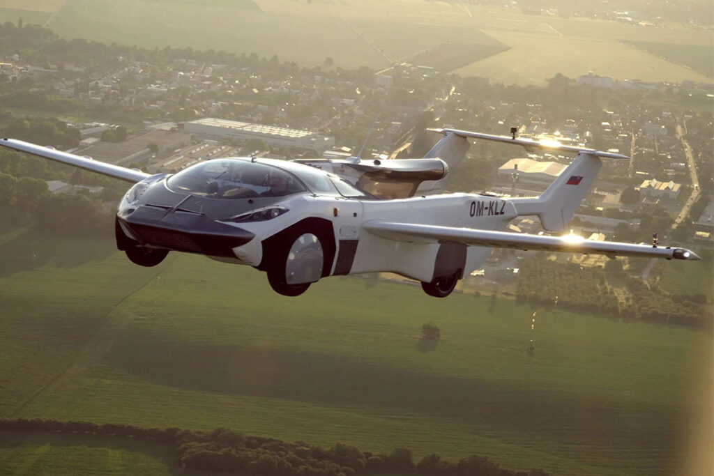 A sleek white flying car with extended wings soars above green fields and a town at sunset.